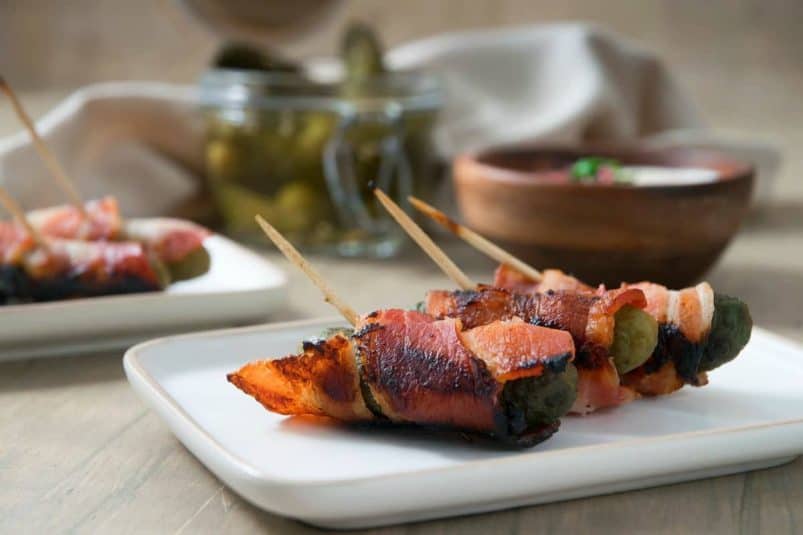 Grilled, bacon wrapped pickles with toothpicks, on a white plate.