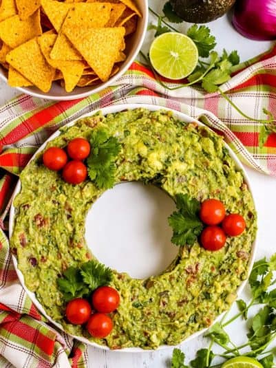 Guacamole garnished with cherry tomatoes and cilantro in the shape of a wreath with a bowl of tortilla chips on the side.