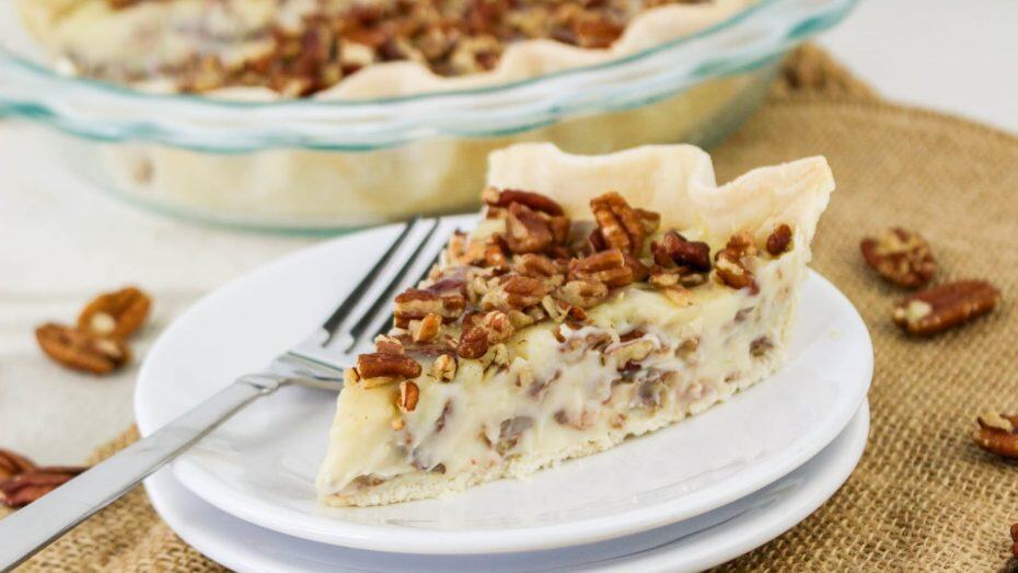 Slice of pecan pie on a white plate with a fork, placed on a burlap surface. The pie has a pastry crust, creamy filling, and is topped with chopped pecans.