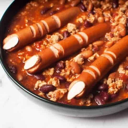 A black bowl with turkey chili and hot dogs in it. The hot dogs have been cut to make them look like witch fingers.