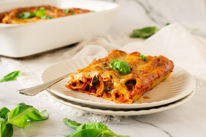 A slice of baked spinach cannelloni on a plate with a fork, garnished with basil, sits invitingly beside a baking dish brimming with deliciousness. In the background, baked spinach cannelloni adds its own charm to the scene, promising an irresistible Italian feast.