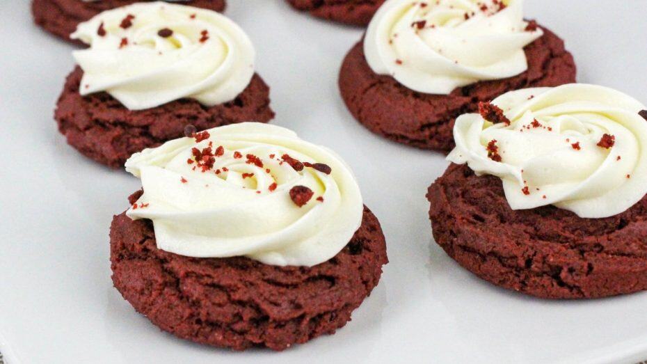 Red velvet cookies topped with swirls of white frosting and red sprinkles on a white surface.