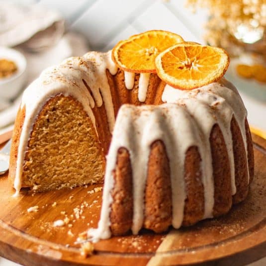 A chai bundt spice cake with a slice cut out, revealing its moist, spiced interior, topped with cream cheese frosting and dried orange slices, presented on a wooden cake stand.