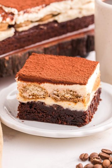 A square slice of tiramisu brownies rests on a white plate, featuring layers of chocolate, cream, and a dusting of cocoa powder.