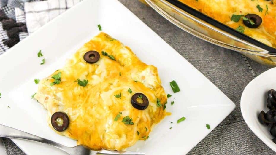 A plate of chicken taquito topped with melted cheese, black olive slices, and chopped parsley. A fork is placed on the plate, and a dish of olives is partially visible in the background.