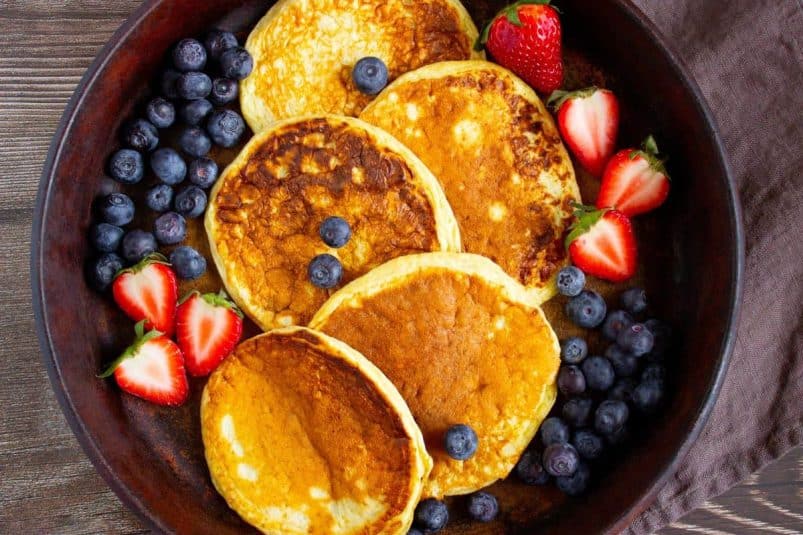 A pan with five high protein cottage cheese pancakes surrounded by blueberries and halved strawberries on a wooden table.