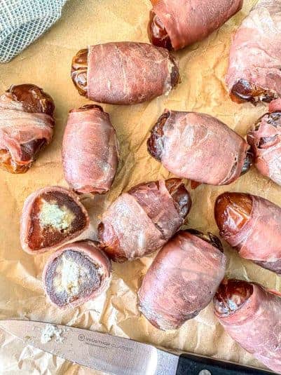 Dates wrapped in prosciutto are scattered on parchment paper. One date is cut open, revealing a creamy filling. A knife rests on the paper nearby.