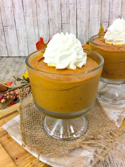 two small trifle dishes filled with pumpkin mousse and topped with whipped cream on a burlap placemat.