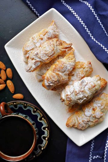 A white rectangle plate with almond croissants, a cup of coffee with a saucer and whoel almonds next to the plate.