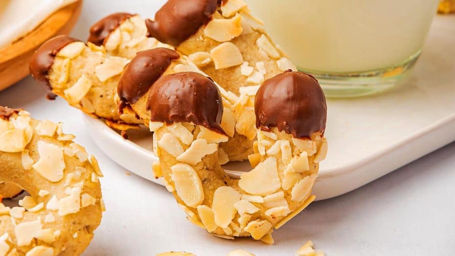 A plate of crescent-shaped pastries dusted with powdered sugar, filled with a dark jam or fruit mixture, placed on a wooden surface. 