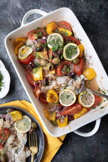 Baked flounder fillets topped with sliced tomatoes, bell peppers, red onions, olives, and lemon slices, garnished with herbs and spices, in a white baking dish.