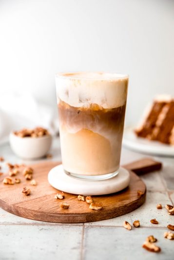 Homemade iced carrot cake latte topped with cream cheese cold foam in a clear pint glass on a white coaster on a wooden cheese tray on a grey slate table with pecan pieces scattered around and a slice of frosted carrot layer cake blurred in the background.