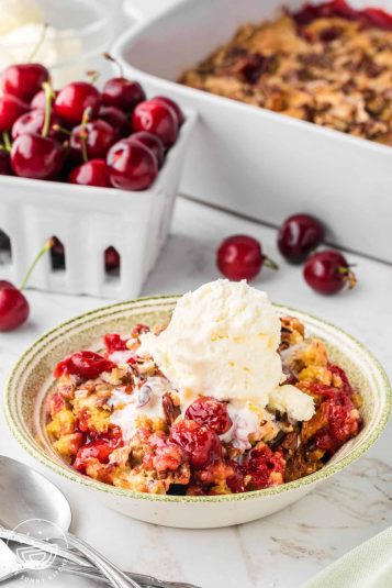 A bowl of cherry dump cake topped with vanilla ice cream, in front of a box of fresh cherries and the full pan of dump cake.