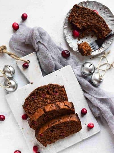 Overhead view of cranberry gingerbread loaf on a grey cutting board, with slice on a plate on a white background.