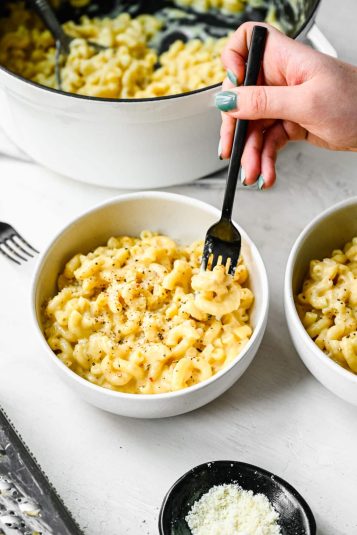 A black fork taking a bite of gouda mac and cheese in a white bowl.