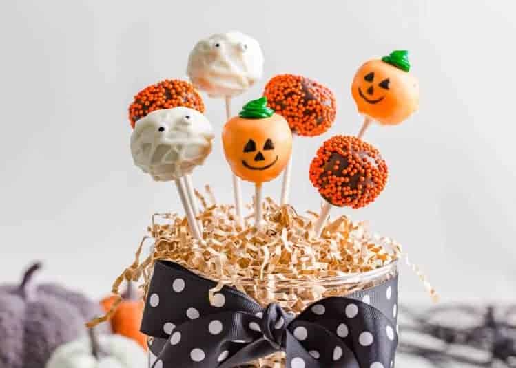 Assorted Halloween cake pops displayed in a jar, decorated as mummies, pumpkins, and chocolate pops with orange sprinkles.