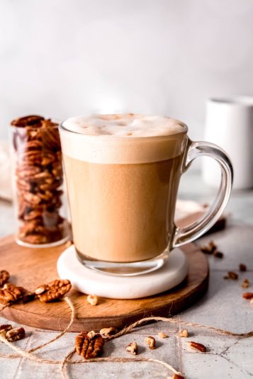 Clear glass mug filled with a creamy homamade pecan crunch oatmilk latte on a white coaster on a wooden tray with a clear collins glass filled with pecan halves in the background and whole pecan halves and pieces scattered around in the foreground.