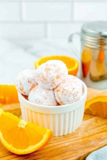 Creamsicle truffles covered in powdered sugar piled high in a bowl with slices of orange near the bowl on a cutting board.