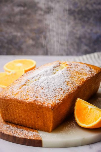 An orange loaf cake on a serving board being sprinkled with icing sugar.