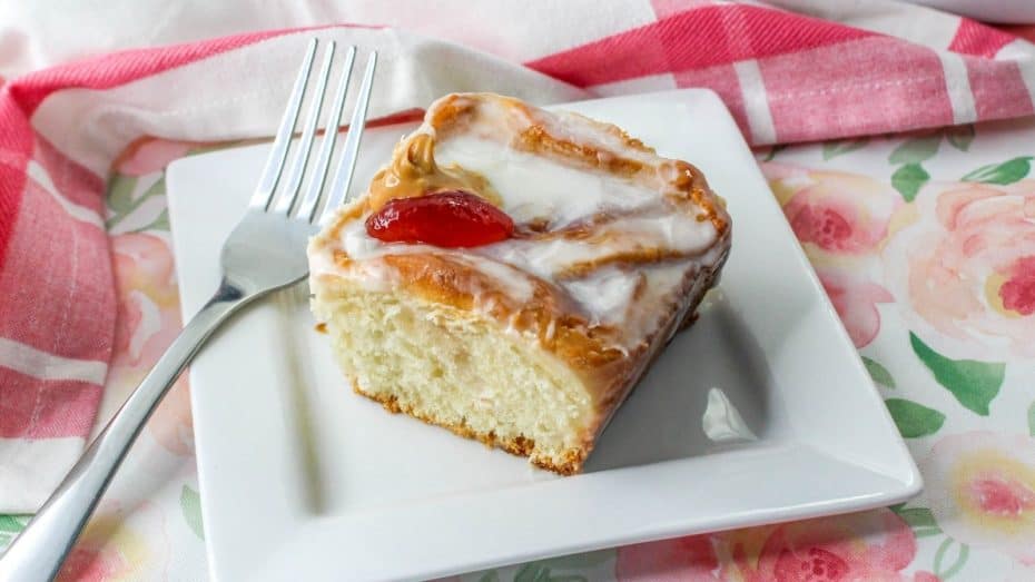 A slice of chocolate cake with a strawberry filling and dark chocolate glaze on a plate. Whole cake in the background, garnished with a halved strawberry. Fork on the side.