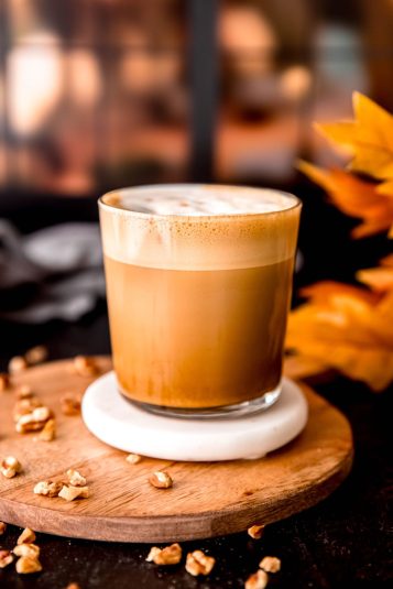 Homemade Starbucks pecan oatmilk cortado in a short, clear glass on a white coaster on a wooden serving tray with pecans sprinkled around and some fall leaves blurred out in the background.