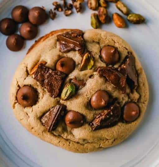 A stack of large, thick cookies with a golden-brown exterior. The cookies are densely packed with visible chunks of milk chocolate, toffee pieces, and green pistachio nuts, showcasing their rich and varied texture.