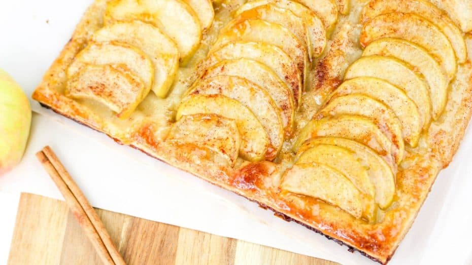 Rectangular apple tart with golden apple slices, a flaky crust, and a light glaze. Two cinnamon sticks lie nearby on a wooden surface.