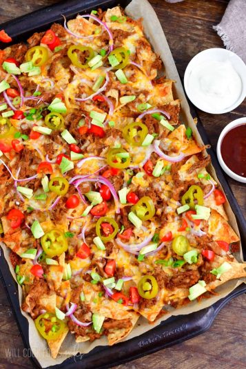 Pulled pork nachos on baking sheet topped with pickled jalapenos, tomato, red onion, and diced avocado.