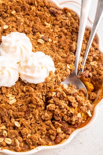 White scalloped pie pan with pumpkin crisp topped with 3 rosettes of whipped cream and 2 silver serving utensils.