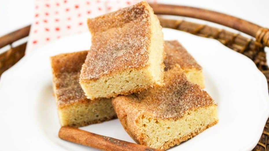 Three cinnamon sugar blondies stacked on a white plate, placed on a woven tray, with a cinnamon stick beside them.