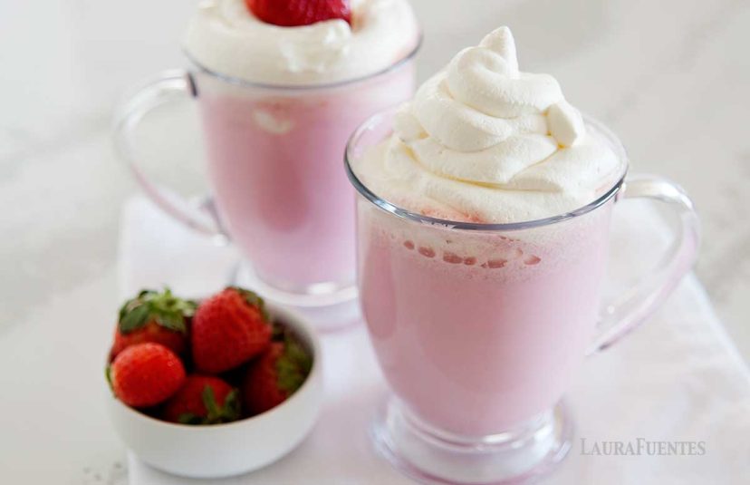 Two clear mugs of pink hot chocolate topped with whipped cream next to a small dish of strawberries.