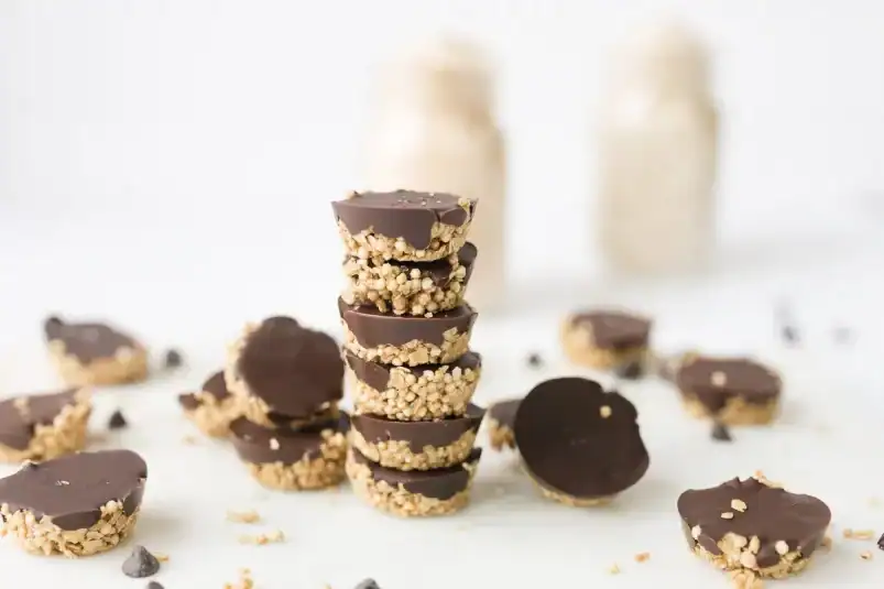 Stacked no-bake SunButter oat cups with puffed quinoa and dark chocolate, surrounded by additional mini cups and scattered chocolate chips on a white surface.