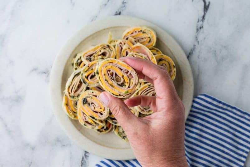 A hand holds a pinwheel-shaped appetizer made of rolled deli meat, cheese, and greens, with more pinwheels arranged on a white plate on a marble surface next to a blue-striped cloth.