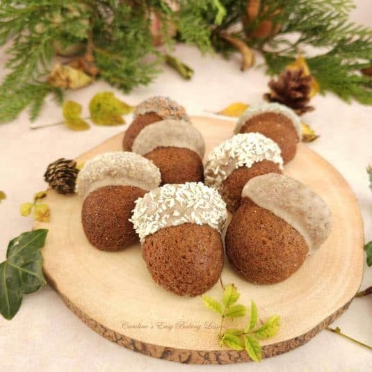 Close shot of mini gingerbread acorn shaped cakes, on round wooden tree-like board, surrounded by Autumn leaves and fircones.