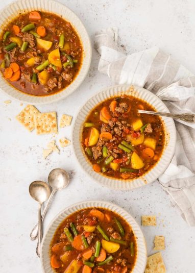 Top shot of a black bowl with instant pot ground beef vegetable soup and a wooden spoon, a white bowl with soup in the top left corner.