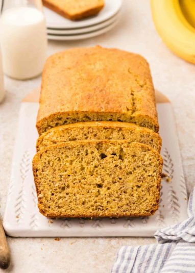 Loaf of sliced banana bread on a white cutting board, accompanied by a glass of milk and a blue-striped cloth. Blurred background includes additional loaf portions and a bunch of bananas.