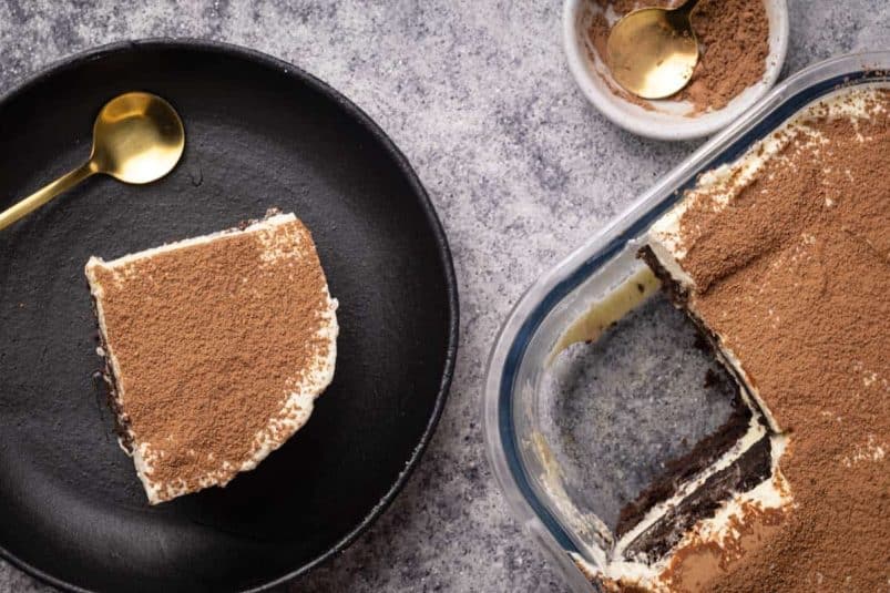 A slice of chocolate tiramisu on a black plate next to the whole dish.