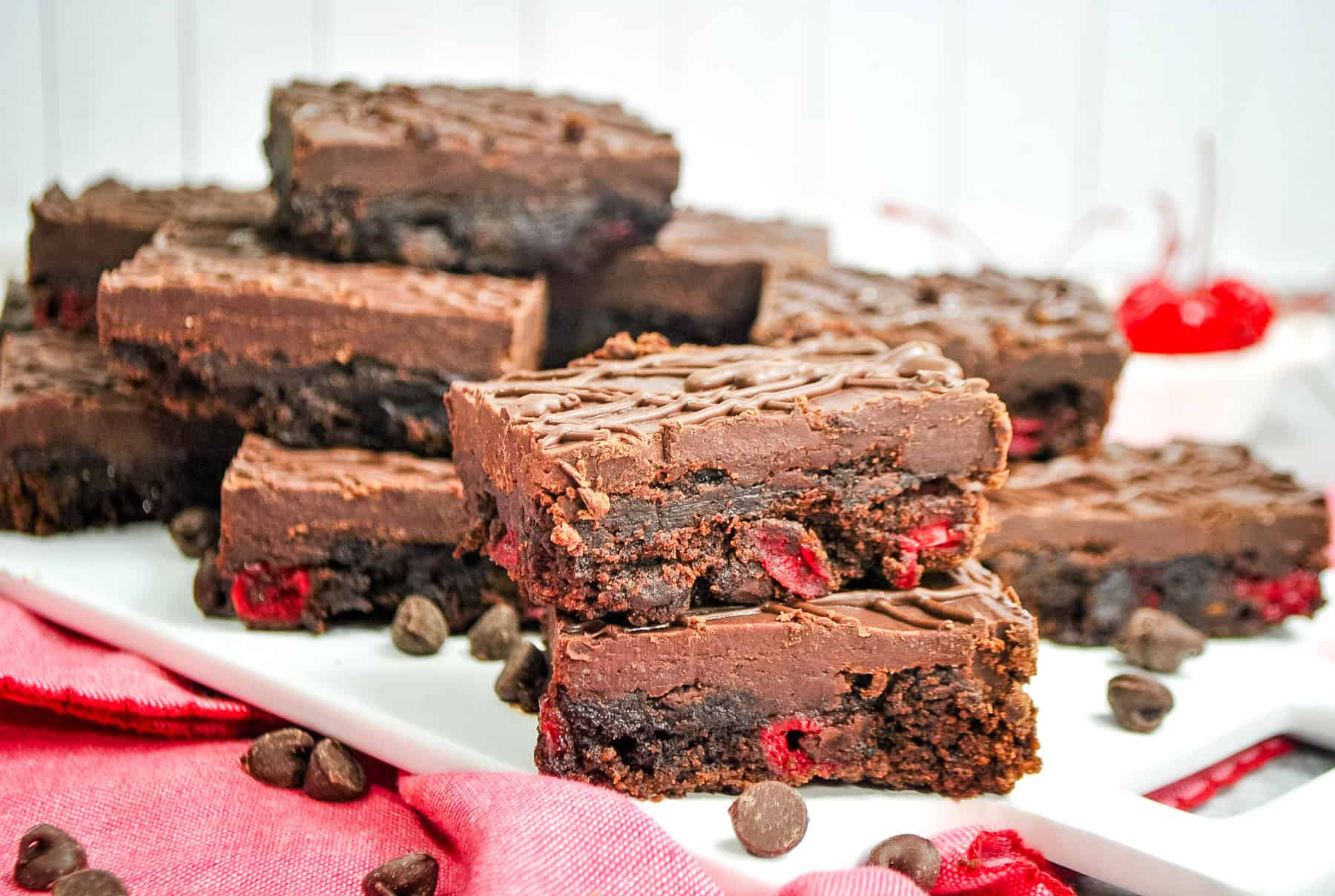 Cherry brownies topped with chocolate ganache on a platter with chocolate chips scattered around.