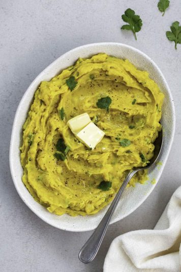 A white bowl filled with curried mashed potatoes, topped with fresh cilantro leaves and butter.