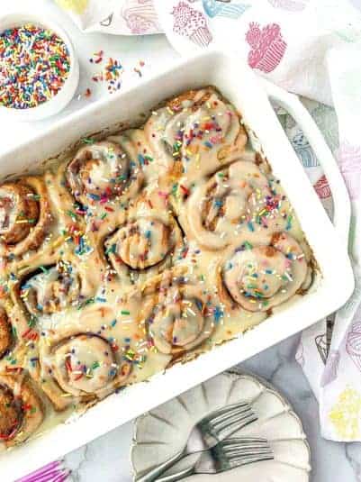 Funfetti Cinnamon Rolls in white pan with rainbow sprinkles nearby.