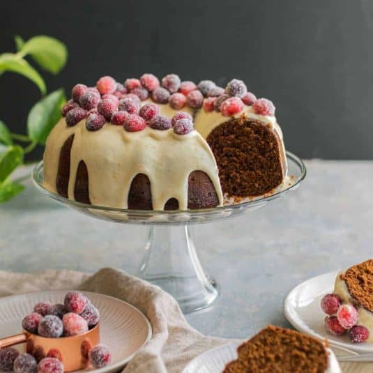 Gingerbread bundt cake elevated on a glass cake stand with frosting and frosted cranberries on top.