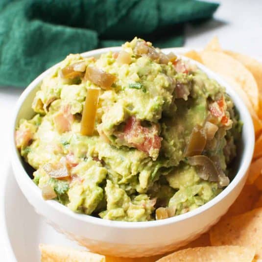 Guacamole with pickled jalepenos in a bowl surrounded by tortilla chips.