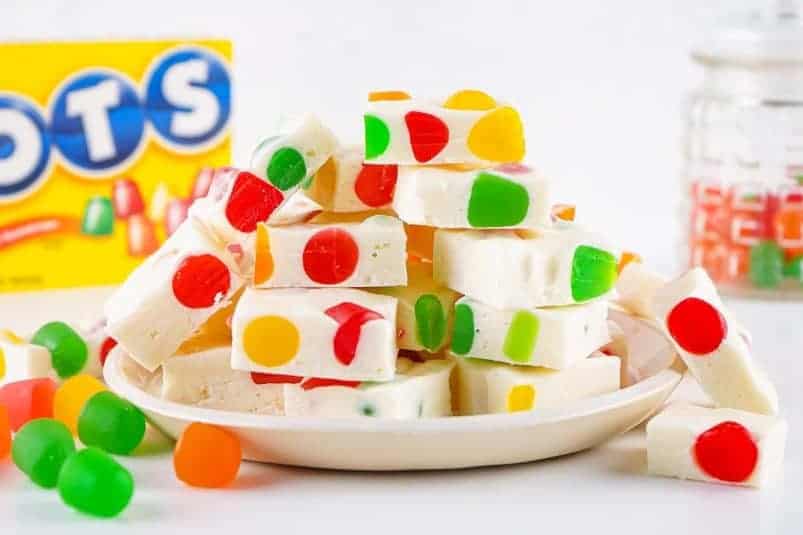 A plate of gumdrop nougat candy squares embedded with colorful Dots candy, with a box of Dots and a glass jar of gumdrops in the background.