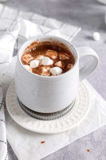 A mug containing oat milk hot cocoa that is sitting on a plate. There are vegan marshmallows floating in the top of the hot cocoa.