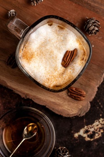 Overhead view of a maple latte topped with a pecan, sitting on a wooden table.