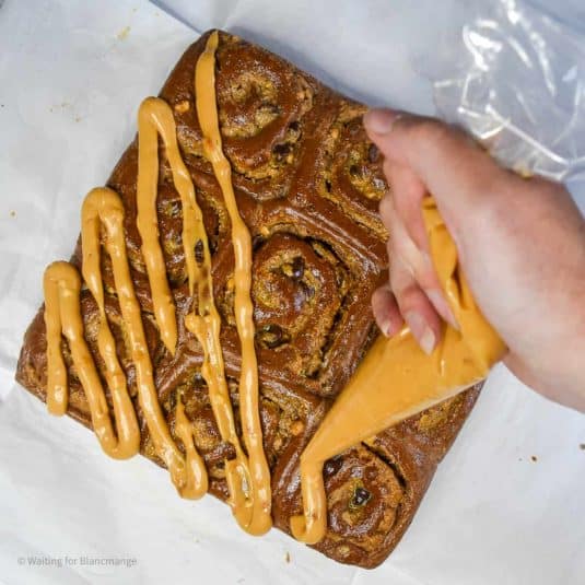 An overhead image of dark chocolate rolls with peanut butter and chocolate chip filling. The rolls have been removed from their pan and are sat on a white countertop on top of parchment paper. There is a hand holding a piping bag over top of them, piping a zig zag of thick peanut frosting.