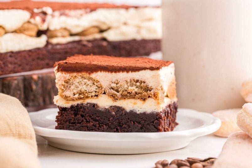 A square slice of tiramisu brownies rests on a white plate, featuring layers of chocolate, cream, and a dusting of cocoa powder.