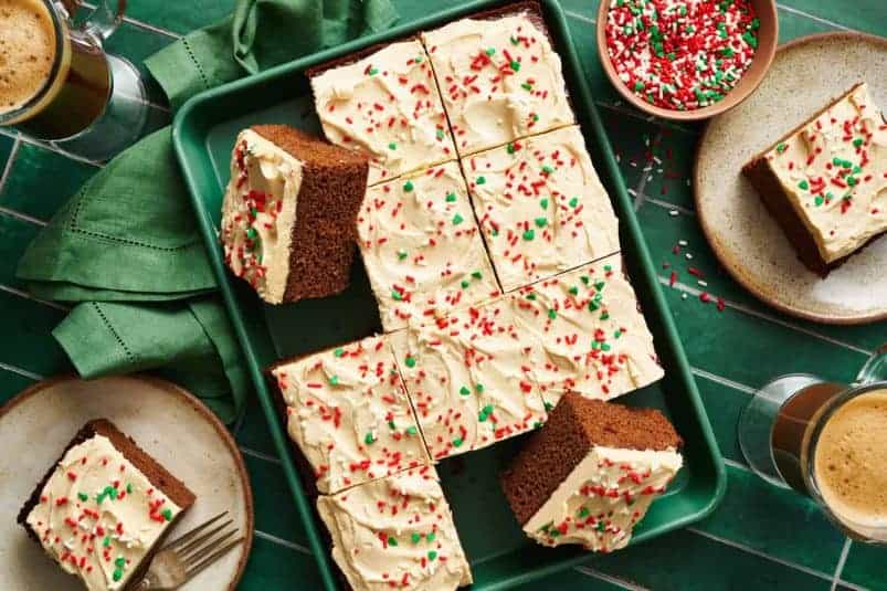 Overhead view of a rectangular gingerbread cake with frosting on top and red and green sprinkles on a green background with a green napkin.