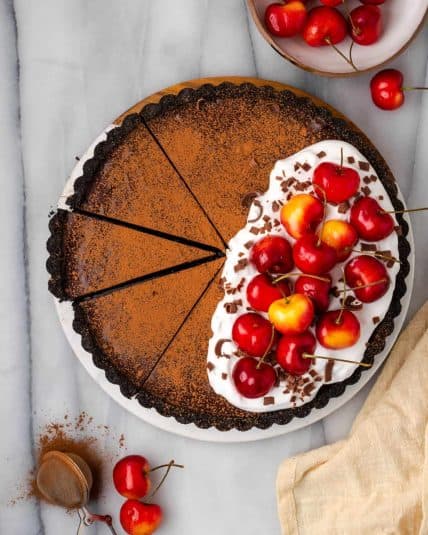 Chocolate ganache tart topped with whipped cream and cherries on a white marble table.