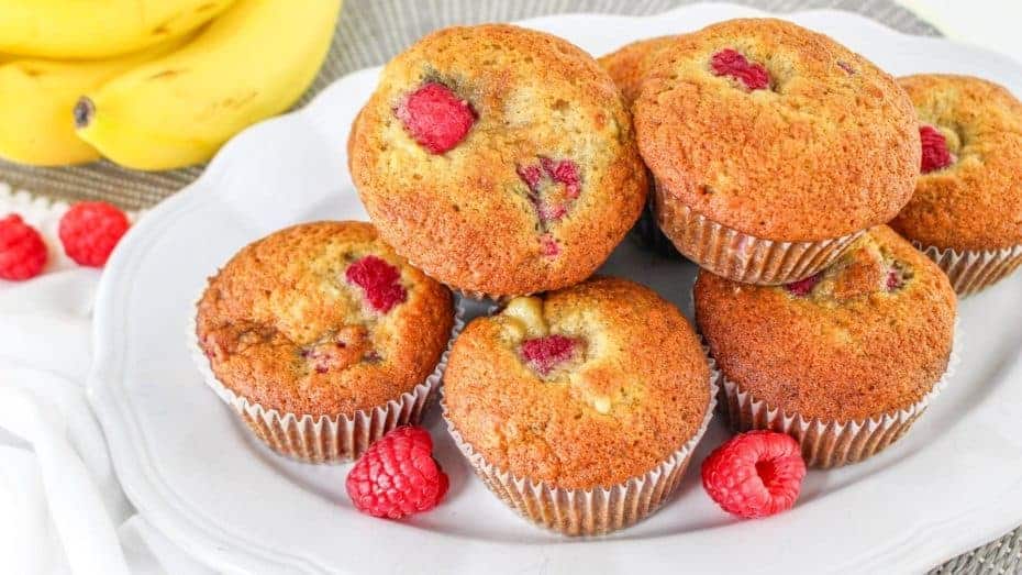 A plate of banana raspberry muffins with raspberries on top and bananas in the background.